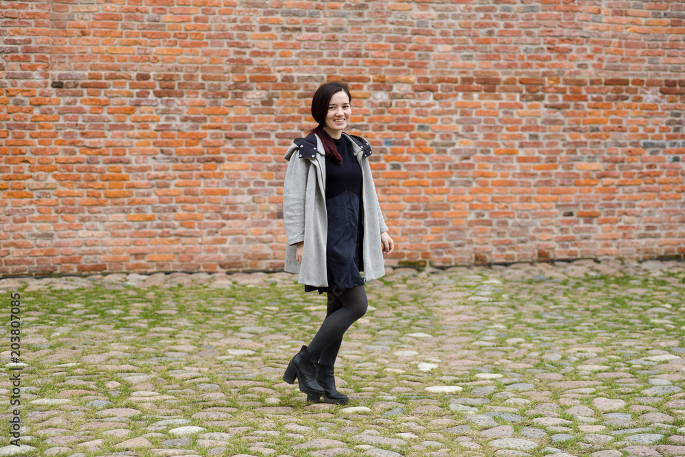 Outdoor portrait of young model of mixed race dressed in black dress and gray coat walking next to the brick wall Outdoor portrait of young model of mixed race dressed in black dress and gray coat walking next to the brick wall