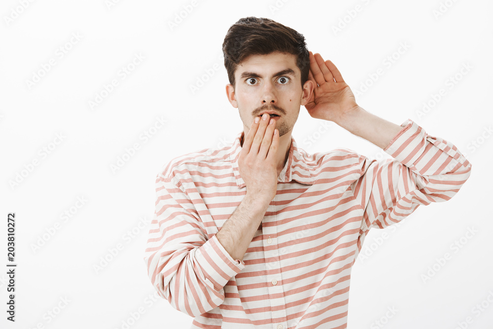 Portrait of shocked intrigued attractive caucasian man in trendy striped shirt, holding hand near ear and mouth, overhearing conversation or eavesdropping, hearing something shocking and interesting Portrait of shocked intrigued attractive caucasian man in trendy striped shirt, holding hand near ear and mouth, overhearing conversation or eavesdropping, hearing something shocking and interesting