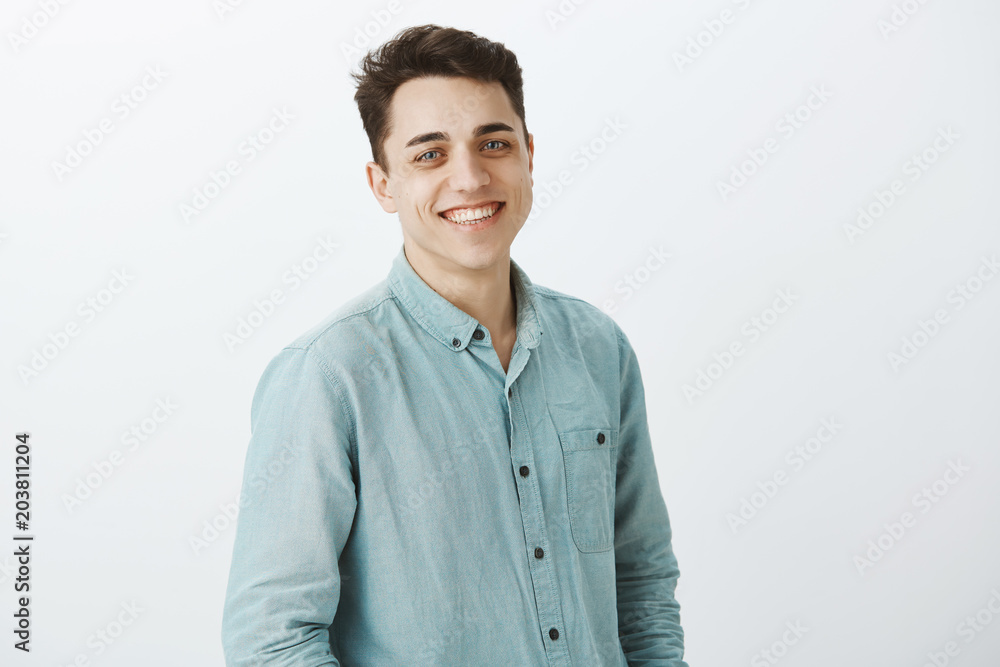 Portrait of good-looking friendly brother standing half-turned over gray background and smiling broadly, talking to customer and giving suggestions of working with company, posing for advertisement Portrait of good-looking friendly brother standing half-turned over gray background and smiling broadly, talking to customer and giving suggestions of working with company, posing for advertisement
