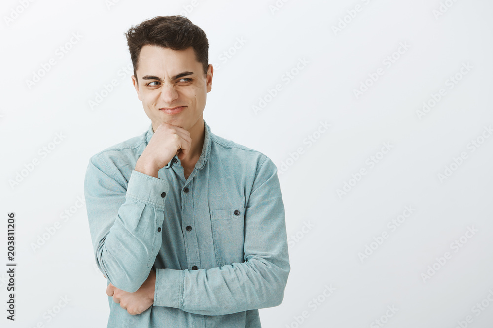 Portrait of attractive male villain in casual clothes, holding hand on chin and looking right with smirk and nasty thoughts, having some bad intentions and making decision over gray background Portrait of attractive male villain in casual clothes, holding hand on chin and looking right with smirk and nasty thoughts, having some bad intentions and making decision over gray background