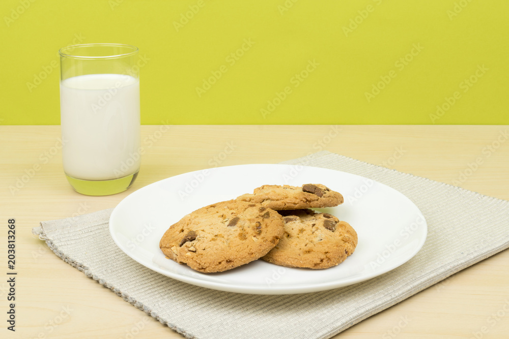 a glass of milk, three pieces of chocolate chip cookies in a white round dish on the wooden table. green background a glass of milk, three pieces of chocolate chip cookies in a white round dish on the wooden table. green background