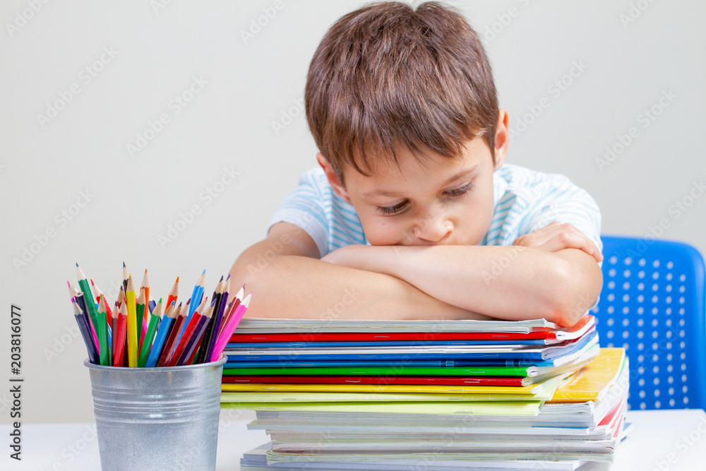 Upset schoolboy sitting at desk with pile of school books and notebooks Upset schoolboy sitting at desk with pile of school books and notebooks
