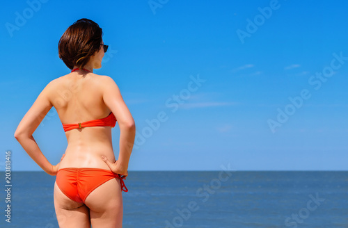 Beautiful woman in orange swimsuit stands looking at the ocean. View from behind at young female in bright bikini standing on the shore and watching the sea. Beach leisure concept