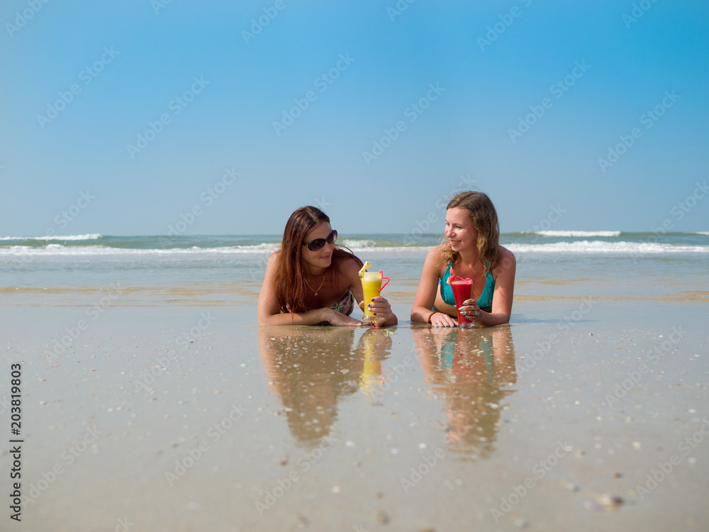 Two white girls with a glass of fruit juice in their hands are lying in the sand on the beach Two white girls with a glass of fruit juice in their hands are lying in the sand on the beach