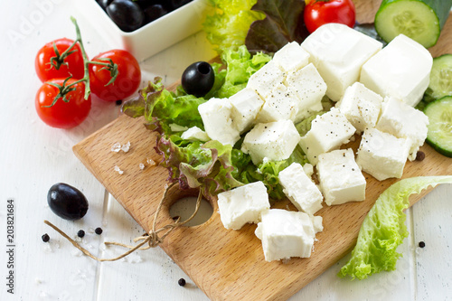 Feta cheese and black olives, cooking qreek salad with fresh vegetables on a white wooden table.