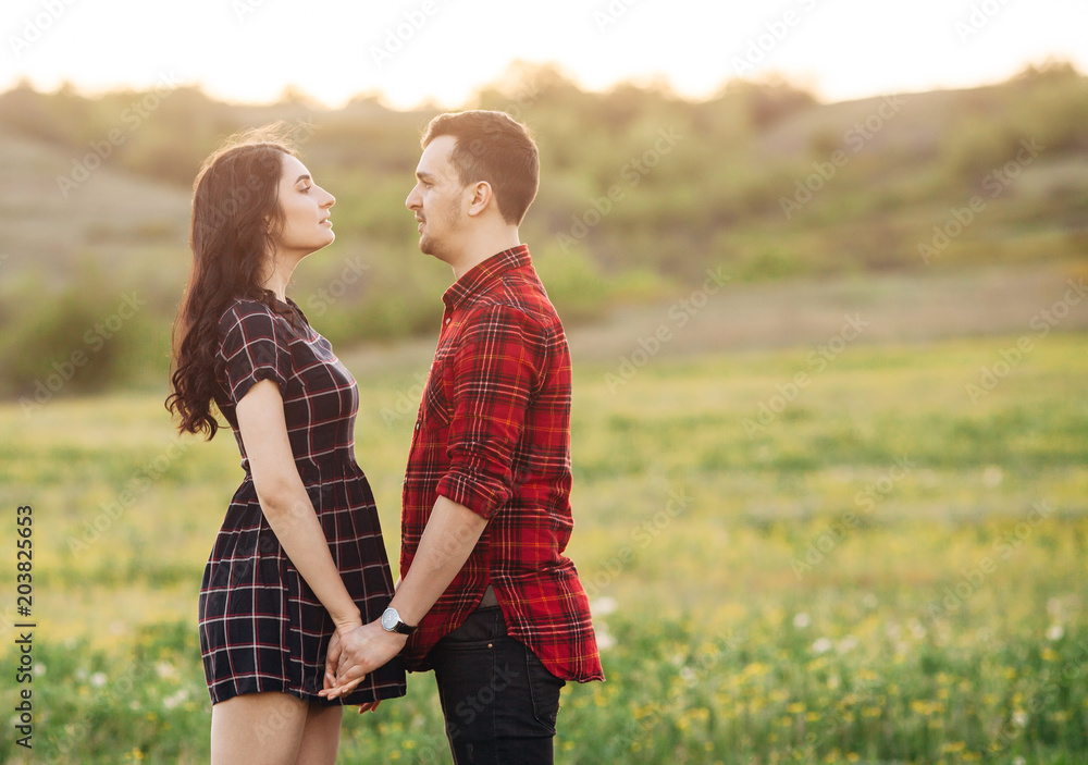 Beautiful couple male and female stand holding hands at the green blooming field. Look to each other. Outdoor Beautiful couple male and female stand holding hands at the green blooming field. Look to each other. Outdoor