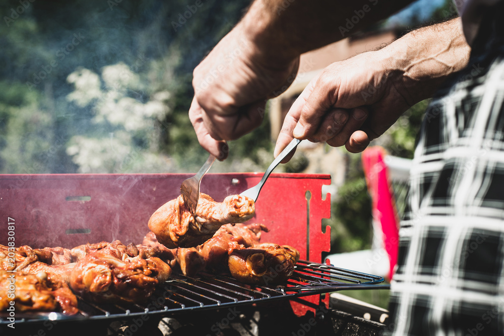 Fototapeta premium Pulling the chicken on the grill. The man pulls the finished chicken legs on the grill. Spending time together with the family at the grill. Social meetings, friends. Red gril with kindling and coal.