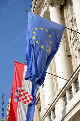 Flaga Unii Europejskiej oraz Chorwacji wiszą razem na scianie budynku, na zewnątrz, powiewają na wietrze, w tle niebieskie niebo