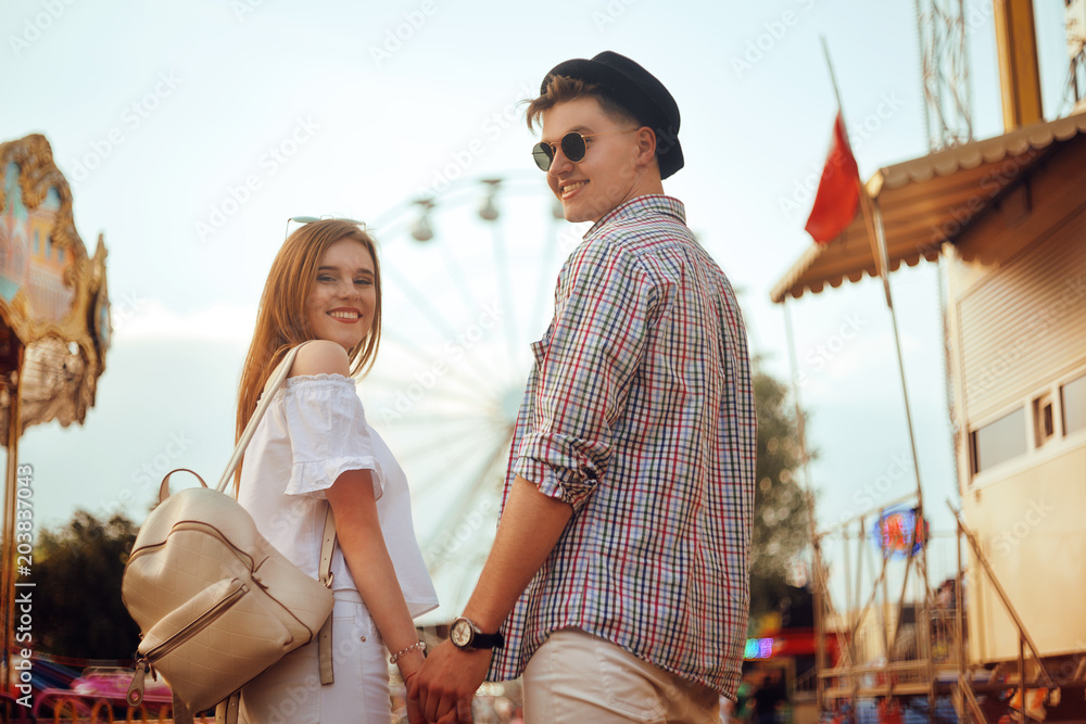 Naklejka premium Beautiful, young couple having fun at an amusement park. Couple Dating Relaxation Love Theme Park Concept. Couple posing together on the background of a ferris wheel. Tourists have fun, smile