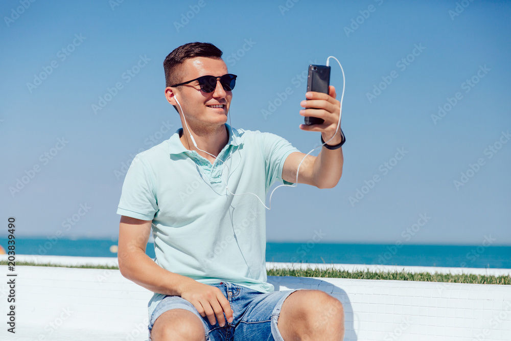 Joyful guy in sunglasses and headset takes a selfie on mobile phone, while sitting on the bench against the sea, wearing summer clothes.