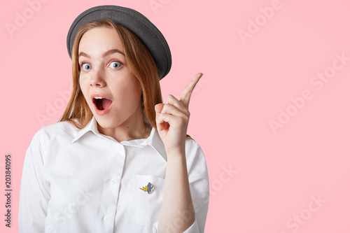 Surprised beautiful European female keeps fore finger raised, wears elegant shirt and hat, gets good idea to put in life, isolated over pink background with copy space for your promotional content photo
