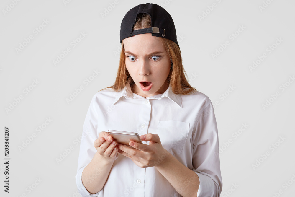 Horizontal shot of teenage girl in stilish cap and shirt, holds smart phone, recieves unexpected message on smart phone, isolated over white background. People, technology and human reaction Horizontal shot of teenage girl in stilish cap and shirt, holds smart phone, recieves unexpected message on smart phone, isolated over white background. People, technology and human reaction