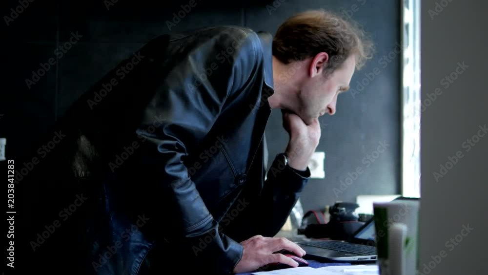 Adult man with black leather coat working with laptop and standing near window at home before he leaving Adult man with black leather coat working with laptop and standing near window at home before he leaving