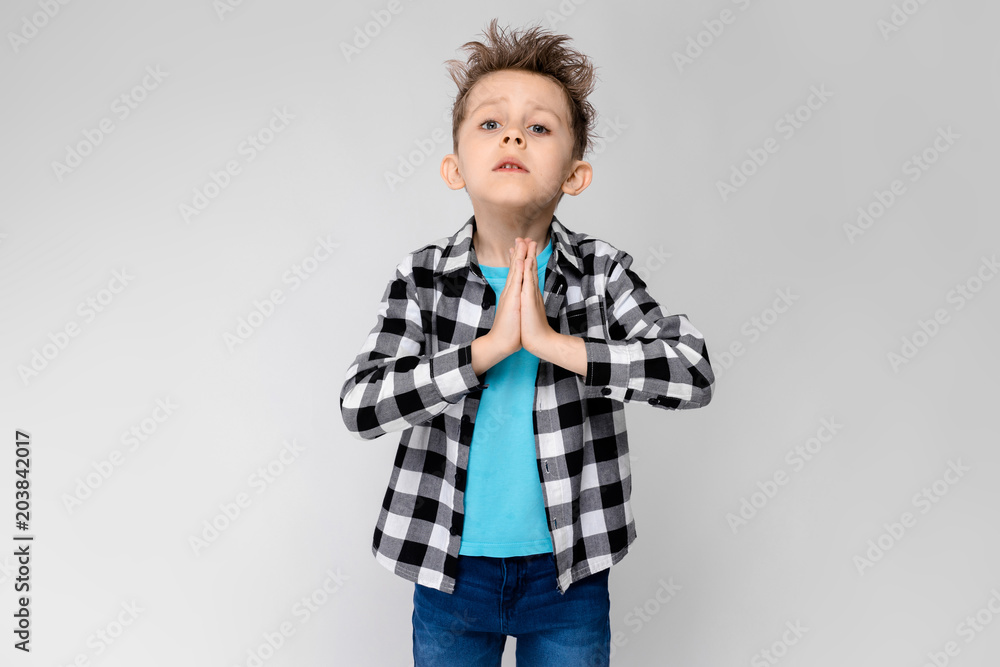A handsome boy in a plaid shirt, blue shirt and jeans stands on a gray background. The boy folded his arms over his chest. The boy folded his palms against his chest A handsome boy in a plaid shirt, blue shirt and jeans stands on a gray background. The boy folded his arms over his chest. The boy folded his palms against his chest
