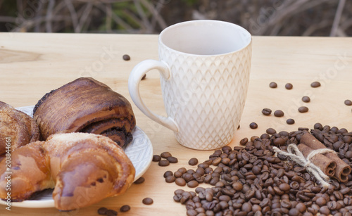 white cup with coffee on a wooden background with rolls on a plate and coffee beans with cinnamon