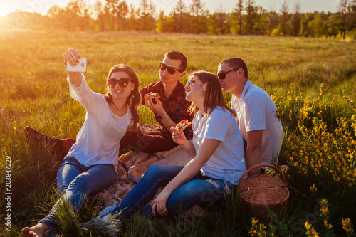 Friends eating pizza outside and taking selfie. Women and men having picnic at sunset. Fast food concept.