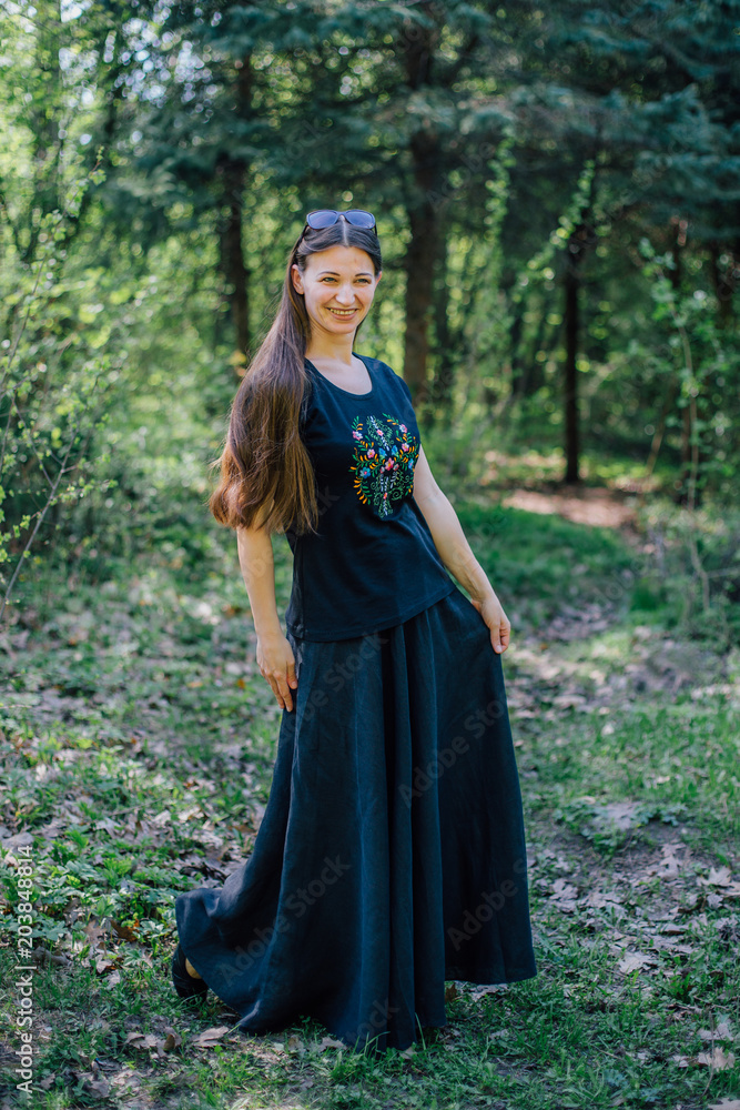 Smiling girl in long black dress with embroidery walks through the woods