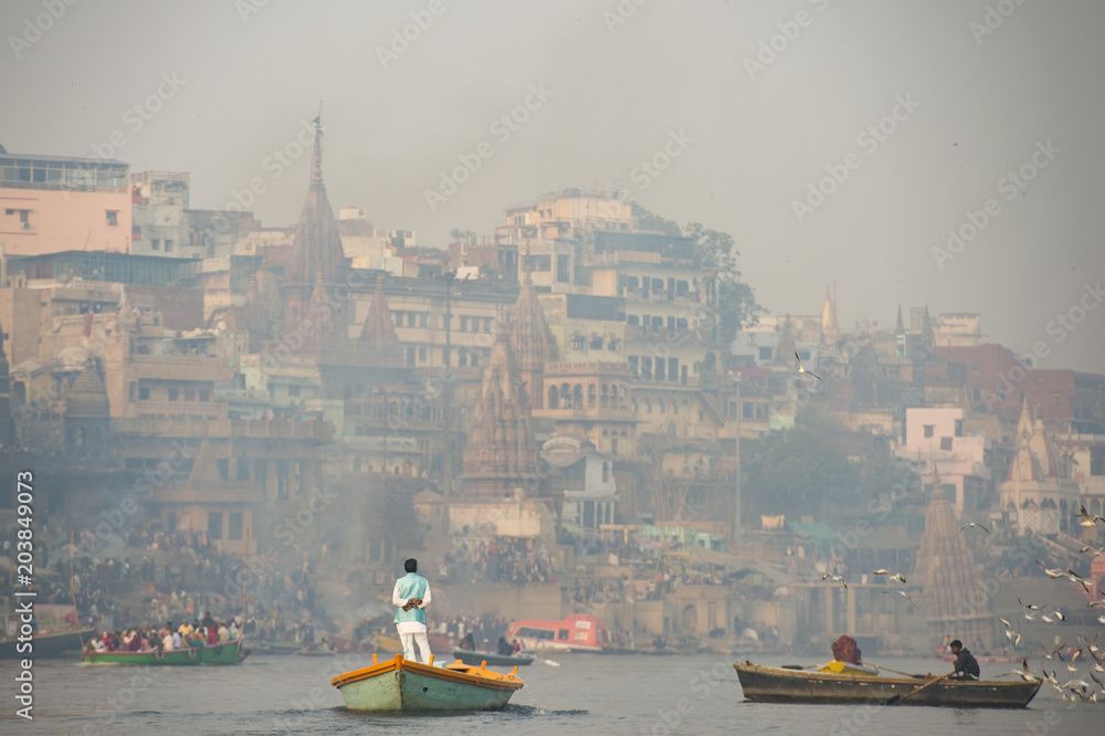 A man is sailing on his wooden boat in the sacred river Ganges in the city of Varanasi at sunset. Blurred Manikarnika ghat on background.