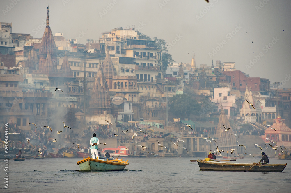 A man is sailing on his wooden boat in the sacred river Ganges in the city of Varanasi at sunset. Blurred Manikarnika ghat on background.
