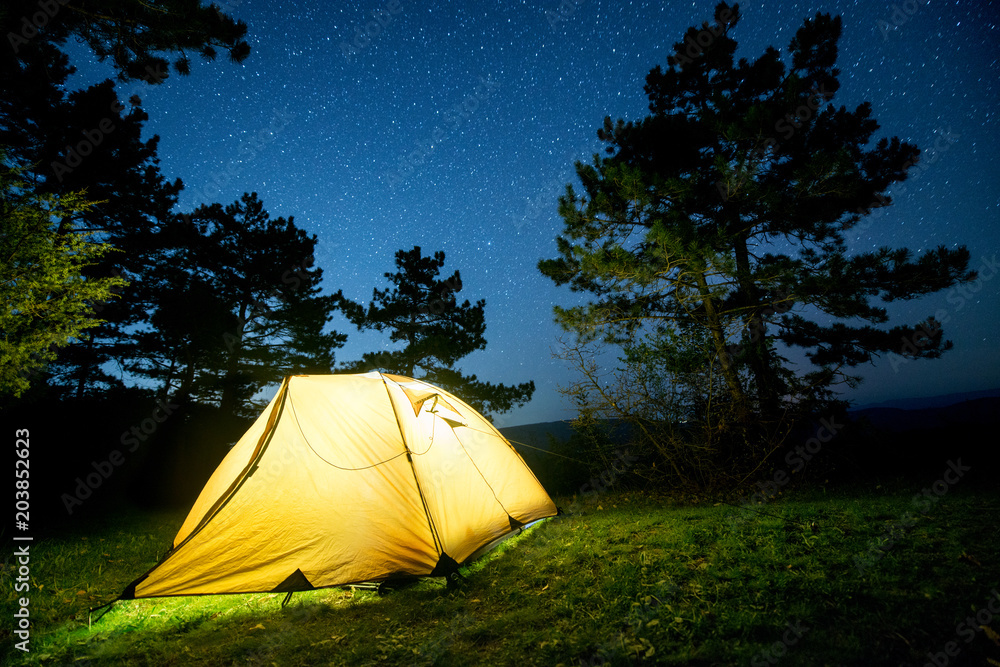 Glowing camping tent in the night mountain forest under a starry sky