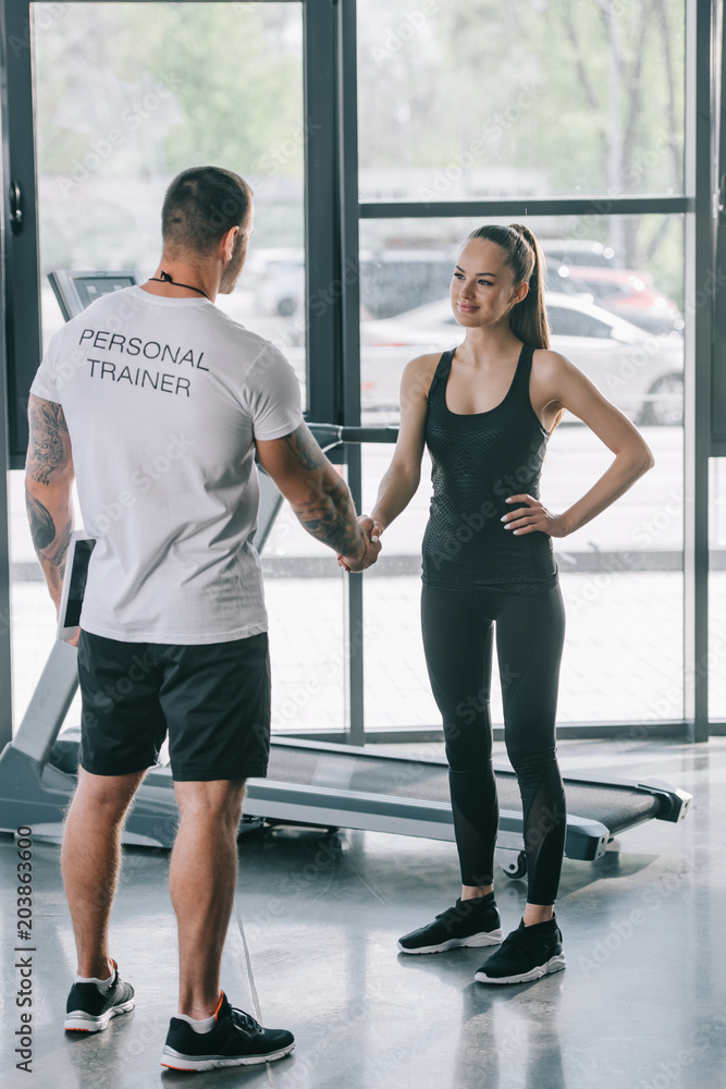 rear view of male personal trainer and young sportswoman shaking hands at gym