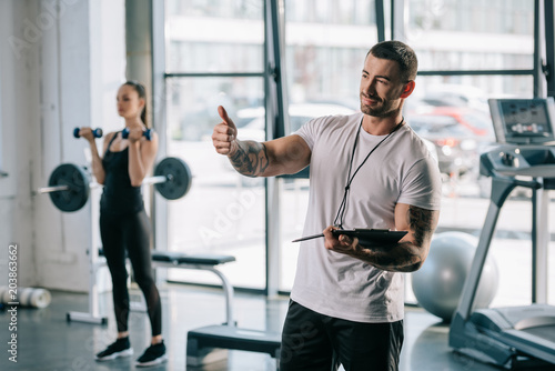 personal trainer doing thumb up gesture and young sportswoman exercising with dumbbells behind at gym