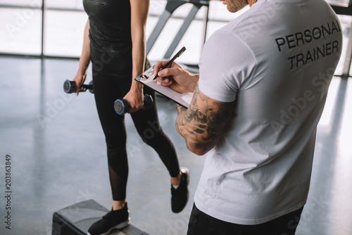 Fototapeta Naklejka Na Ścianę i Meble -  cropped image of male personal trainer writing in clipboard and young sportswoman doing step aerobics exercise with dumbbells at gym