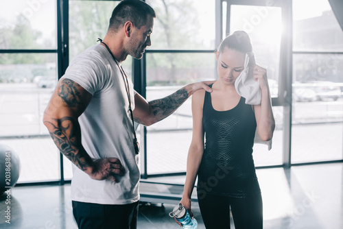 male personal trainer cheering up young sportswoman with bottle of water wiping face with towel at gym