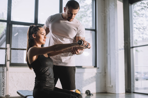 male personal trainer helping sportswoman to do exercises with dumbbells at gym