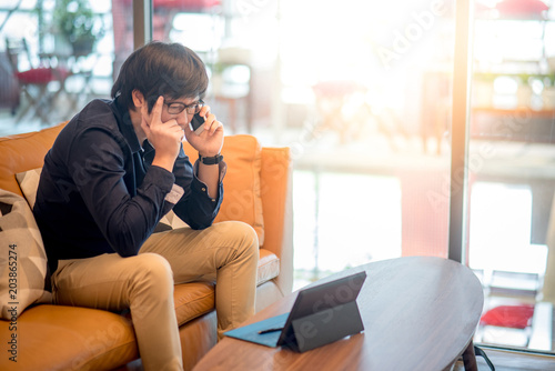 Young Asian businessman feeling stressed and frustrated while calling on smartphone working with digital tablet in living space. Business problem concept
