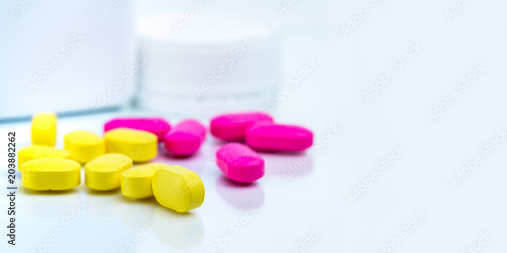 Selective focus on yellow ovoid rectangular tablet pills on blurred background of pink tablet pills and medicine bottle with copy space. Pharmacy or drugstore concept. Pharmaceutical industry. Selective focus on yellow ovoid rectangular tablet pills on blurred background of pink tablet pills and medicine bottle with copy space. Pharmacy or drugstore concept. Pharmaceutical industry.