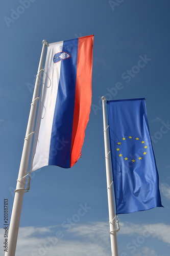 Flaga Słowenii i Unii Europejskiej wiszą obok siebie na drzewcach, powiewają na wietrze, w tle błękitne niebo