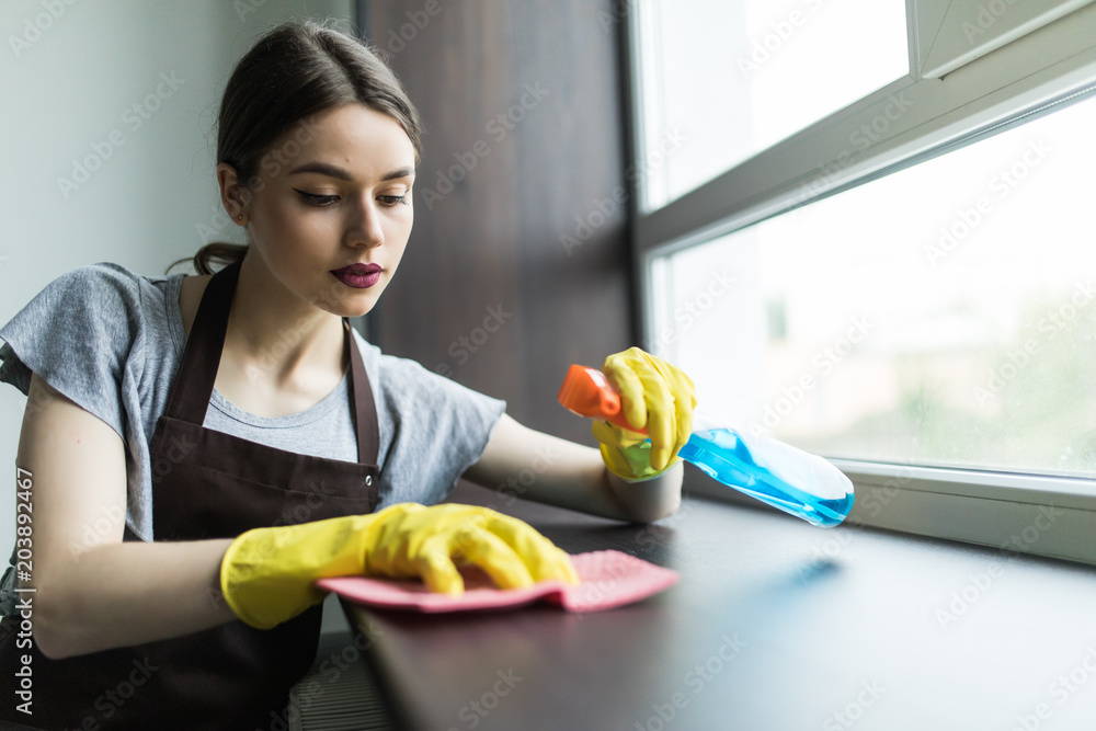 Young woman in yellow gloves holding rag and window cleaner in hands near the window indoors Young woman in yellow gloves holding rag and window cleaner in hands near the window indoors