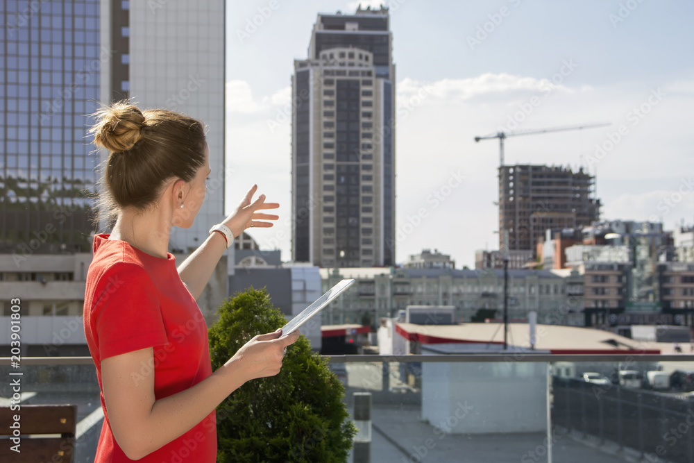 A young woman in a red business dress looks at high-rise buildings, holds a tablet in her hands and points to something in the distance.