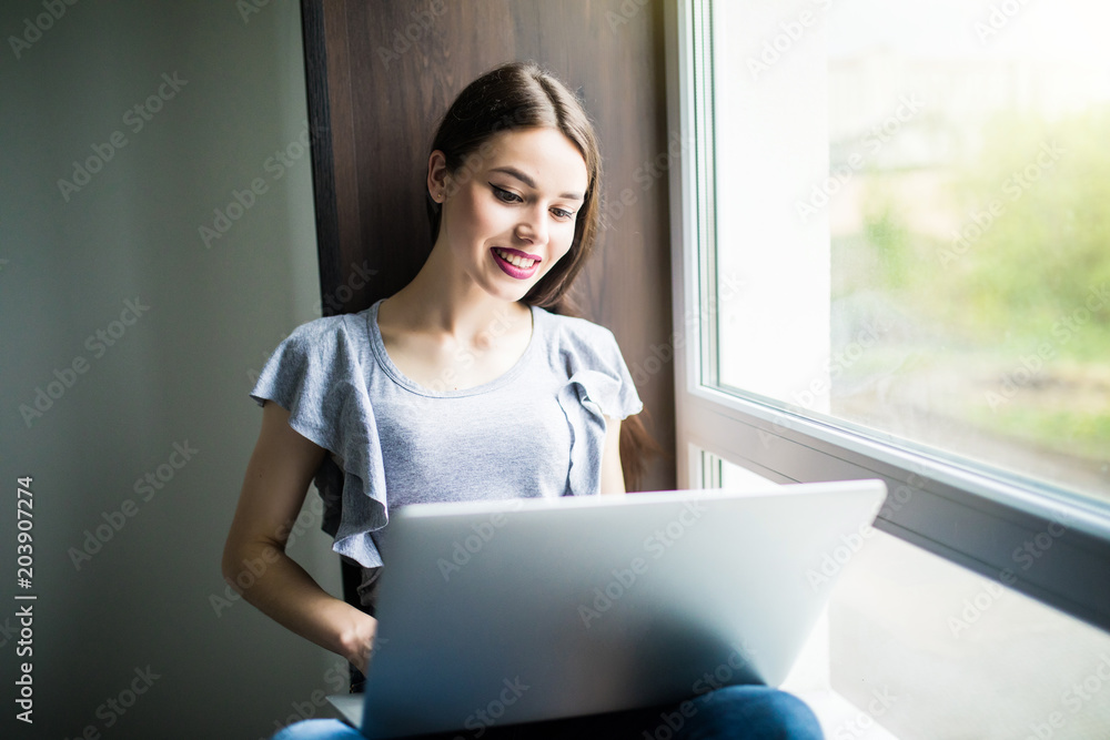 Young woman in t-shirt sitting on windowsill while using laptop computer Young woman in t-shirt sitting on windowsill while using laptop computer