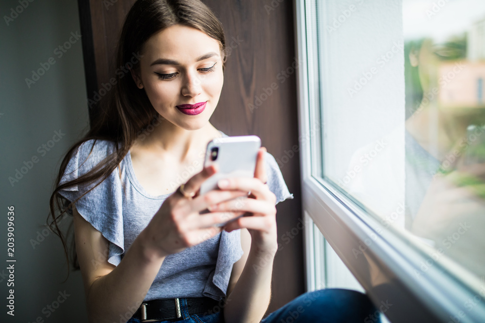 Bored woman watches something on the Internet in her mobile phone and smiling while sitting on windowsill Bored woman watches something on the Internet in her mobile phone and smiling while sitting on windowsill