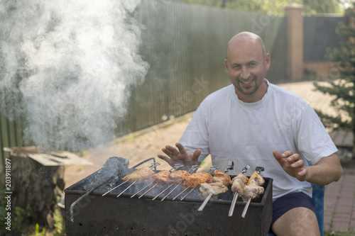 A man with a beard near the fire and the grill fry meat and sausages. A young man fries a barbecue on a grill on a summer day.