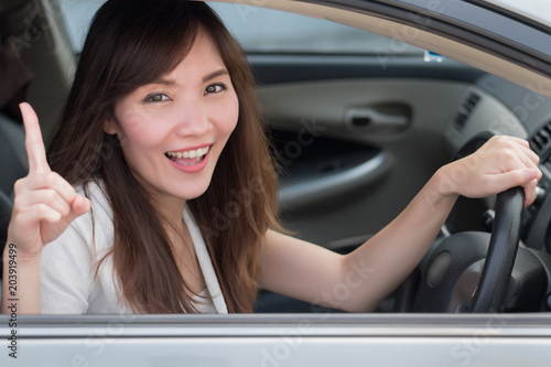 Wallpaper Mural smiling woman driver driving and pointing up one finger; portrait of happy, joyful asian woman driver in left hand driving style pointing up number one gesture; 30s adult asian Chinese woman model Torontodigital.ca