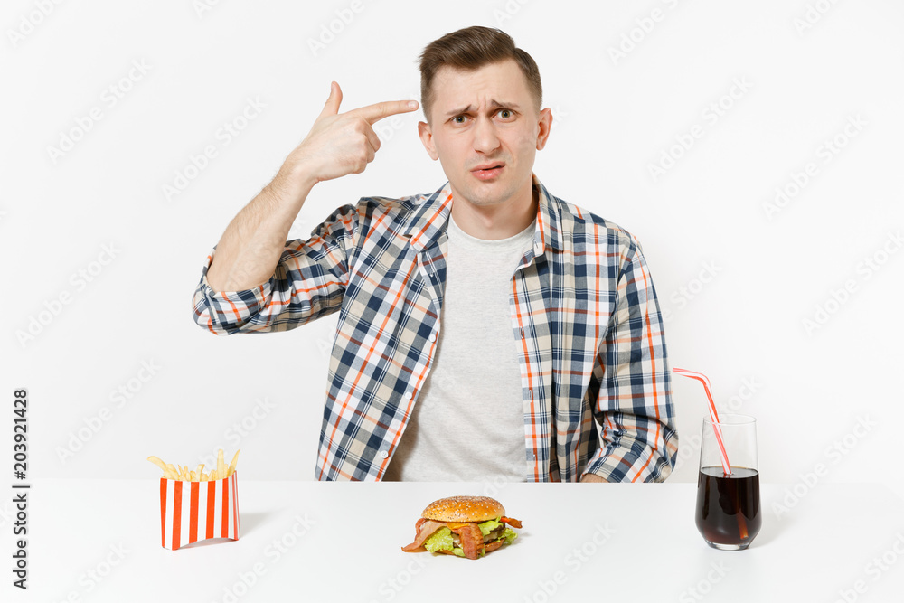 Man point fingers to head as if he shoot himself, sits at table with burger french fries cola in glass isolated on white background. Proper nutrition or American classic fast food. Area to copy space. Man point fingers to head as if he shoot himself, sits at table with burger french fries cola in glass isolated on white background. Proper nutrition or American classic fast food. Area to copy space.