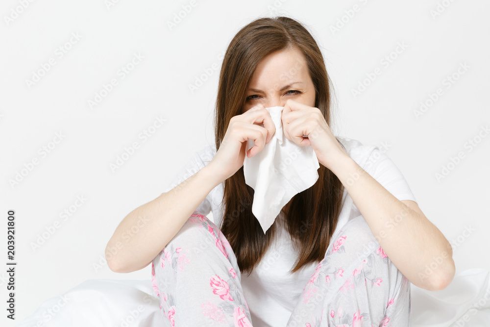 Young sickness tired brunette woman in pajamas sitting on bed isolated on white background. Female feeling bad, blowing her nose into white napkin. Cold in the head. Copy space for advertisement. Young sickness tired brunette woman in pajamas sitting on bed isolated on white background. Female feeling bad, blowing her nose into white napkin. Cold in the head. Copy space for advertisement.