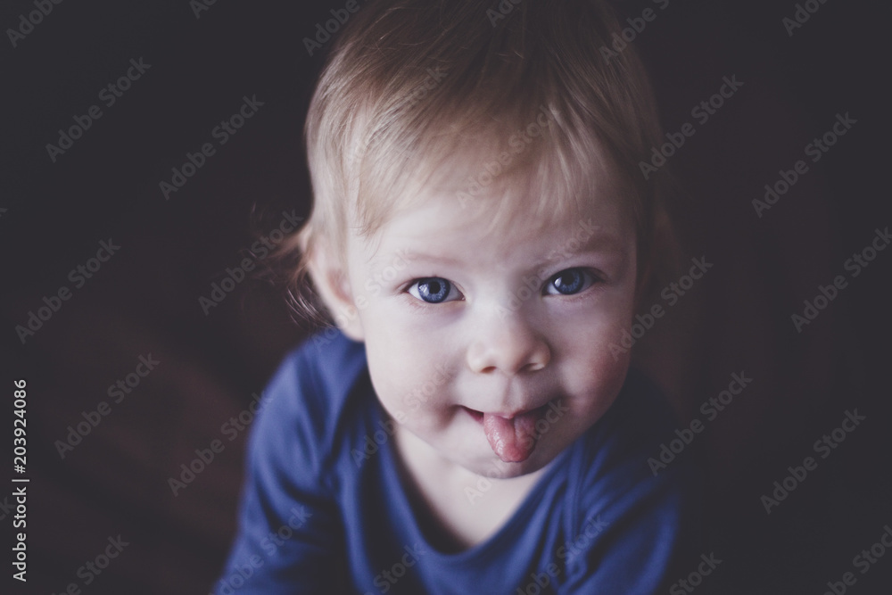 Cute little blond kid with blue eyes smiles slyly and shows his tongue. Dark background. Close up. Imitation of film grain. Cute little blond kid with blue eyes smiles slyly and shows his tongue. Dark background. Close up. Imitation of film grain.