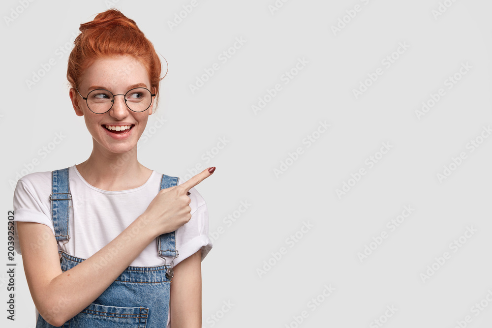 Horizontal shot of happy female student with pleasant smile, being glad to advertise something, points at blank copy space for your promotional text or advertisement. Ginger youngster gestures Horizontal shot of happy female student with pleasant smile, being glad to advertise something, points at blank copy space for your promotional text or advertisement. Ginger youngster gestures