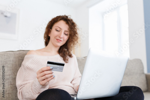 Pretty girl wearing beige casual sweater sits on cozy sofa at home, uses online banking on laptop pc to transfer money from credit card, surfing websites while shopping online