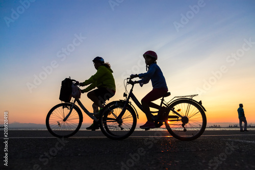 Fototapeta Naklejka Na Ścianę i Meble -  Silhouettes of people enjoying a walk by bicycle the seaside of the town during sunset