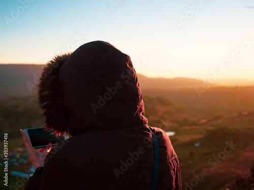travel and photographer concept from backside of woman with hoodie cloth stand and see sunrise from mountain view and use her mobile phone