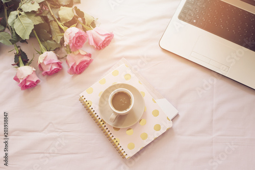 Coffee, old vintage camera in bed on pink sheets. Roses, notebooks and laptop around. Freelance fashion home femininity workspace