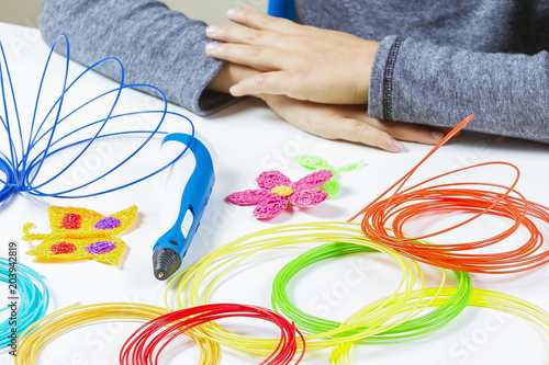 Kid hands and 3d pen, colorful filaments on white desk.