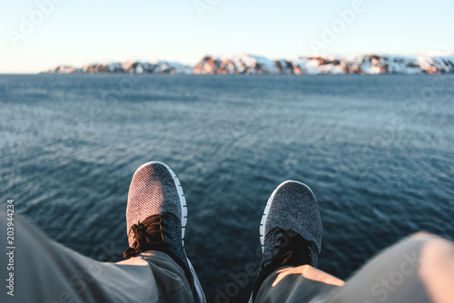 Happy traveler sitting on rock and looks on legs and shoes. POV view on feet on background of ocean and mountains. Adventure vacations outdoor concept