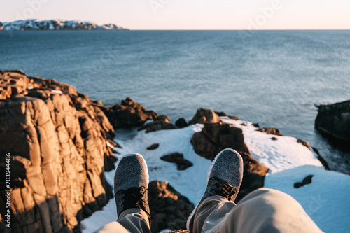 Hipster sitting on cliff in front of breakage above sea and snow mountains. POV view of legs and shoes on background blue water and rocks. Adventure and lifestyle