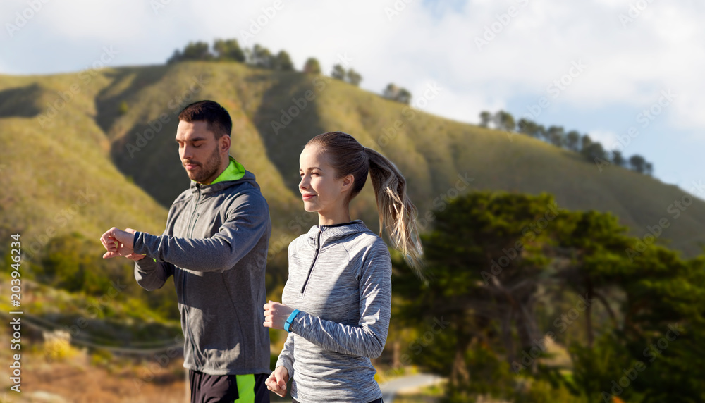 fitness, sport and healthy lifestyle concept - smiling couple with heart-rate watches running over big sur hills background in california